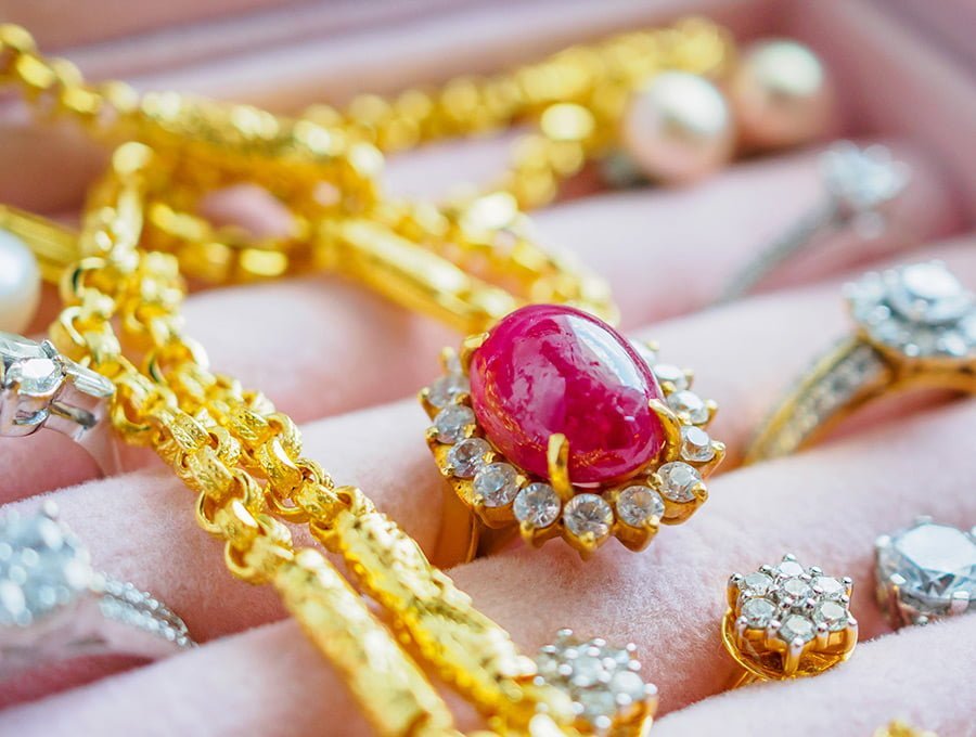Aqui tenemos una cadena de oro de 18 quilates con una gema de color rosa fucsia. Está dentro de un muestrario de joyas junto a varios anillos de oro y brillantes.