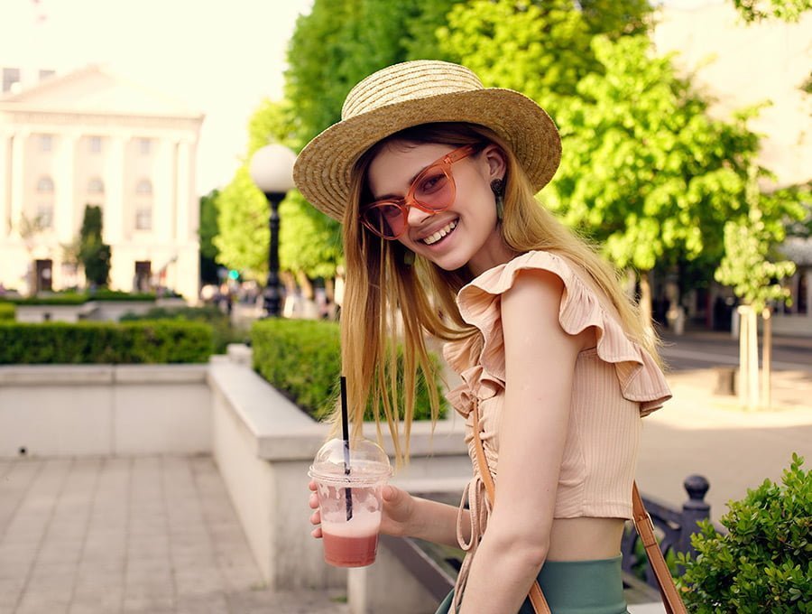 Esta chica con ropa fresca de verano y un sombrero de paja, se está tomando una granizada de frambuesa mientras da un paseo por la ciudad. Está sonriente porque se ha puesto su perfume de Bur preferido.