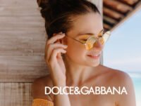 Esta chica está apoyada en la pared de madera de un chiringuito de la playa. Está feliz porque lleva puesta su fragancia D&G preferida, además de sus gafas de sol favoritas a juego con el vestido naranja.