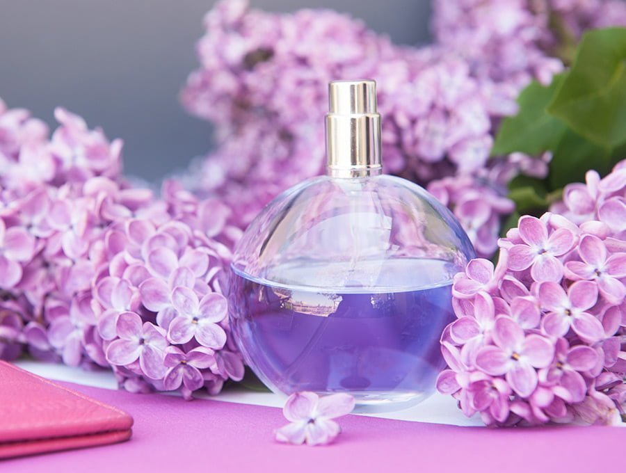 Un bote de perfume con el cristal de color morado, rodeado de flores de violeta. El spray es de color plateado.