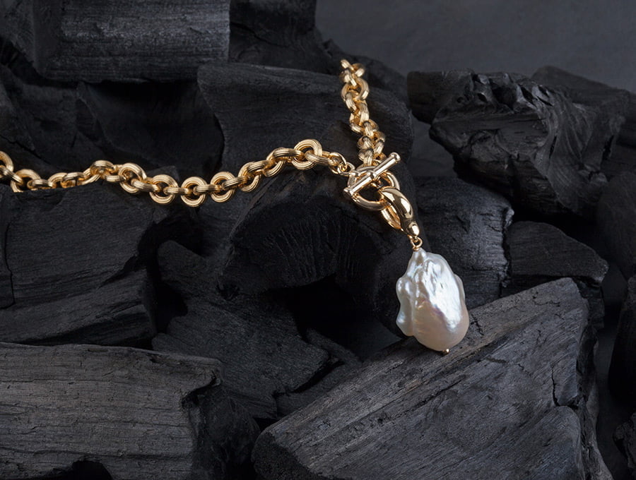 Una cadena de oro hueco con un colgante de perla cultivada de gran tamaño. Está sobre unos trozos de carbón vegetal.