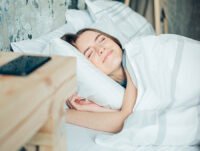Esta chica tiene una cara de felicidad mientras duerme. Está tapada con un edredón nórdico en su cama. En la mesita de noche hay un móvil de color negro.