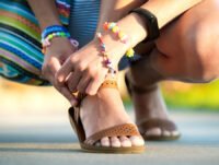 Esta chica joven está agachada abrochándose una de las sandalias. Es verano y lleva un vestido de colores, además de algunas pulseras llamativas a juego con el vestido.