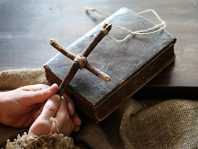 Esta persona está sujetando un colgante con forma de cruz. Está hecho con dos palos de madera cruzados y una cuerda. También hay una biblia encima de la mesa.