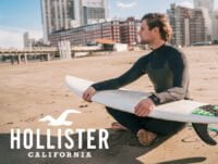 Este surfista está sentado en la arena de la playa. Está mirando al horizonta. Tiene la table de surf cogida encima de sus piernas. Diría que está esperando una buena ola para meterse en el agua y surfearla.