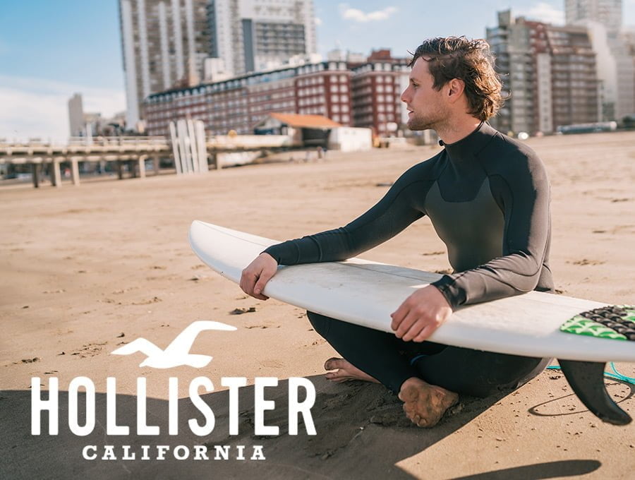 Este surfista está sentado en la arena de la playa. Está mirando al horizonta. Tiene la table de surf cogida encima de sus piernas. Diría que está esperando una buena ola para meterse en el agua y surfearla.