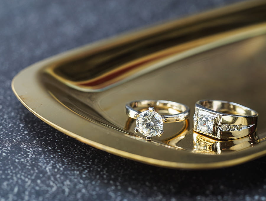 Dos anillos de plata con diamantes simulados engastados. Están sobre una bandeja de metal, encima de la mesa.