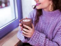 Esta chica con jersey de hilo grueso de color morado, se está tomando un delicioso chocolate caliente junto a la ventana. Lleva unos aretes dorados grandes.