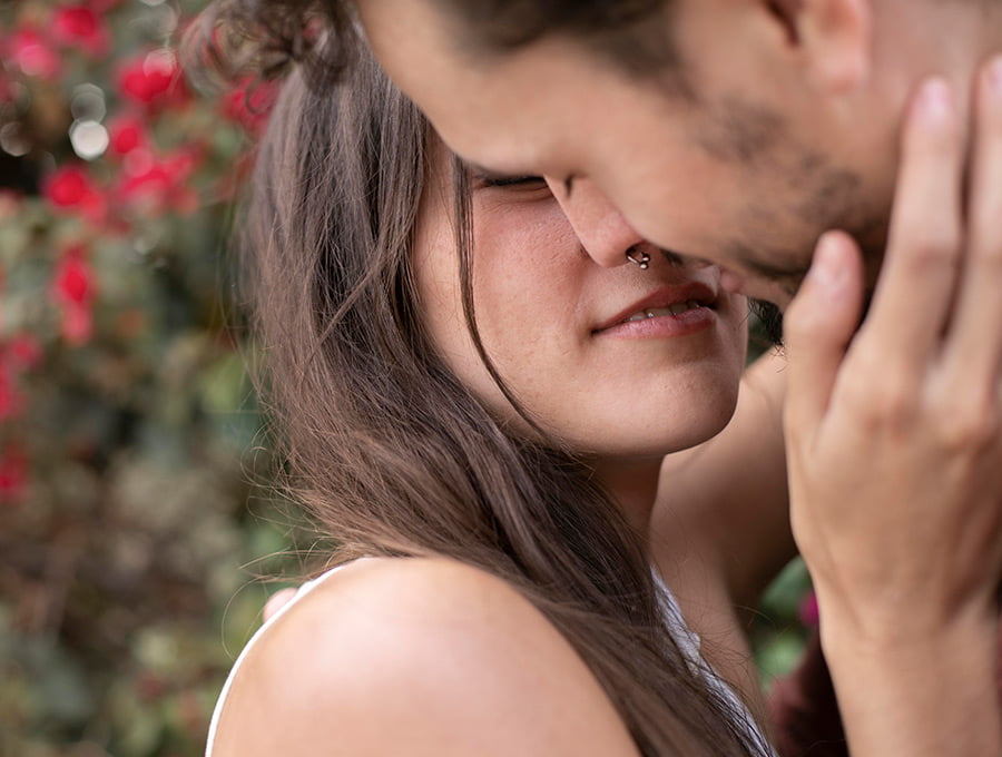Esta pareja de enamorados tiene los labios muy juntos porque se van a dar un beso apasionante. Detrás de ellos hay un rosal con flores rojas muy bonito y grande.