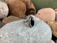 Un anillo con una piedra de onice negro fabricado en acero inoxidable. Está colocado encima de una gran piedra de río.