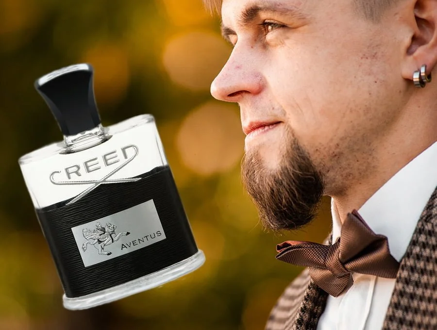 Un hombre trajeado con perilla y un par de pendientes en su oreja. Está mirando un frasco de perfume Creed Aventus.