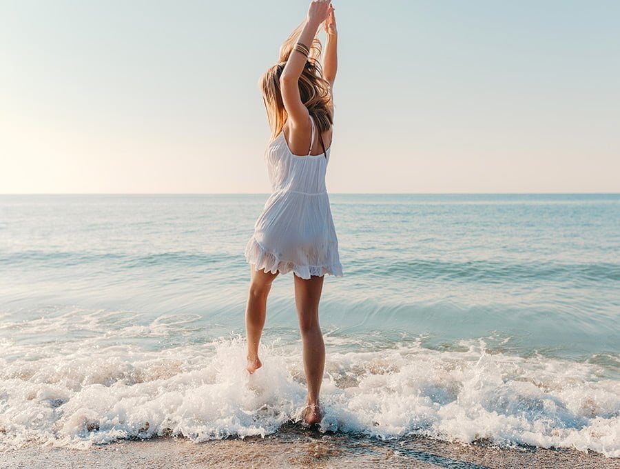 Esta mujer con vestido blanco se va a meter en el agua de la playa. Está feliz.