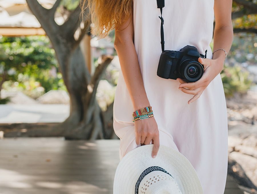 Esta chica lleva puesto un vestido de verano blanco. Tiene una cámara reflex de color negro en su cuello. Además, un bonito sombrero blanco en su mano.