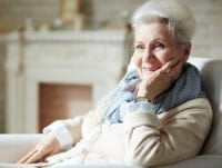 Esta abuela sonriente está sentada en el sillón de su salón. Lleva puesto un anillo. Hay una chimenea al fondo.